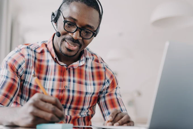 Business professional wearing headset taking notes next to laptop.