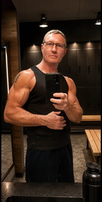 Homme de plus de 60 ans en débardeur noir prenant un selfie dans un vestiaire de salle de sport après sa séance, avec une expression calme et détendue.