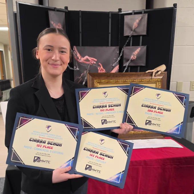 A person is holding four certificates in an indoor setting with display boards in the background.