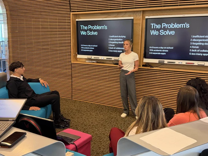 A group of people sitting in a classroom, listening to a person standing at a chalkboard that displays "The Problems We Solve".