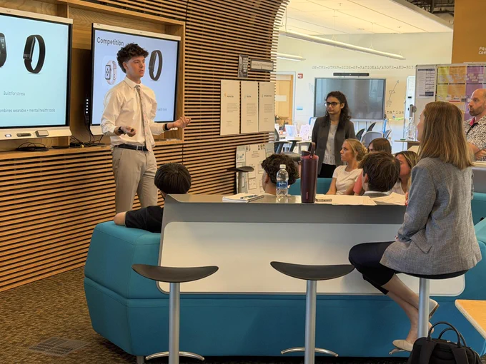 People sitting and listening to a young man presenting with whiteboards and computer screens visible.