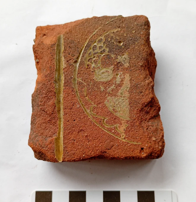 A very worn fragment of a red medieval floor tile, a design outlined in yellow shows a bird bending its neck to its breast. A stylised nest is below the bird.
