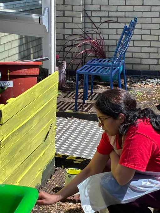 Woman in red shirt painting a wooden planter bright yellow in a sunny outdoor garden area.