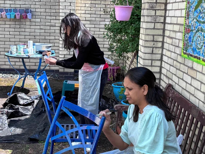 Two people painting blue chairs in a sunlit outdoor garden area with brick walls and potted plants.