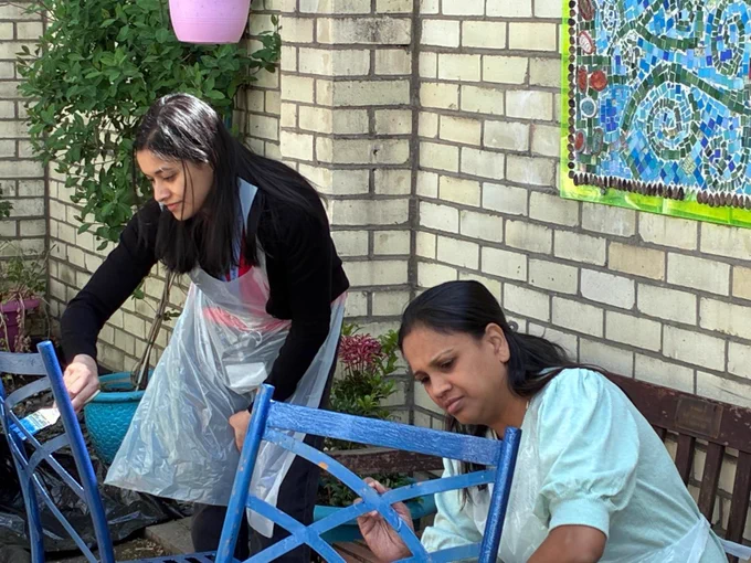 Two people painting blue chairs in a sunlit outdoor garden area with brick walls and potted plants.