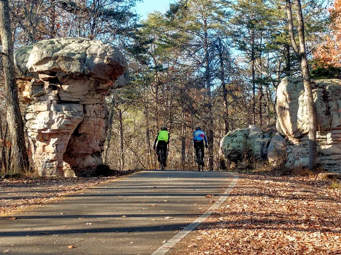 Two people are riding bikes on a road and are passing two unique sandstone formations. The road is lined with trees and brown leaves cover the ground. 