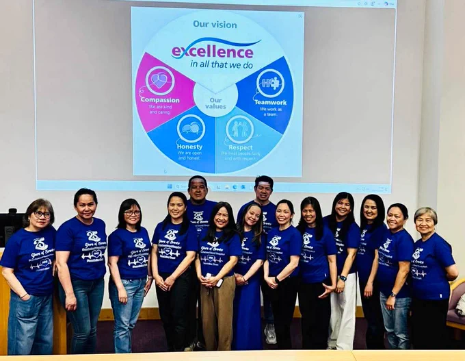 A group of people wearing blue shirts stands in front of a screen displaying a presentation slide. The slide shows a circular graphic with the text "Our vision" at the top and the words "excellence in all that we do" in the center. The circle is divided into sections labeled "Compassion," "Teamwork," "Honesty," and "Respect.