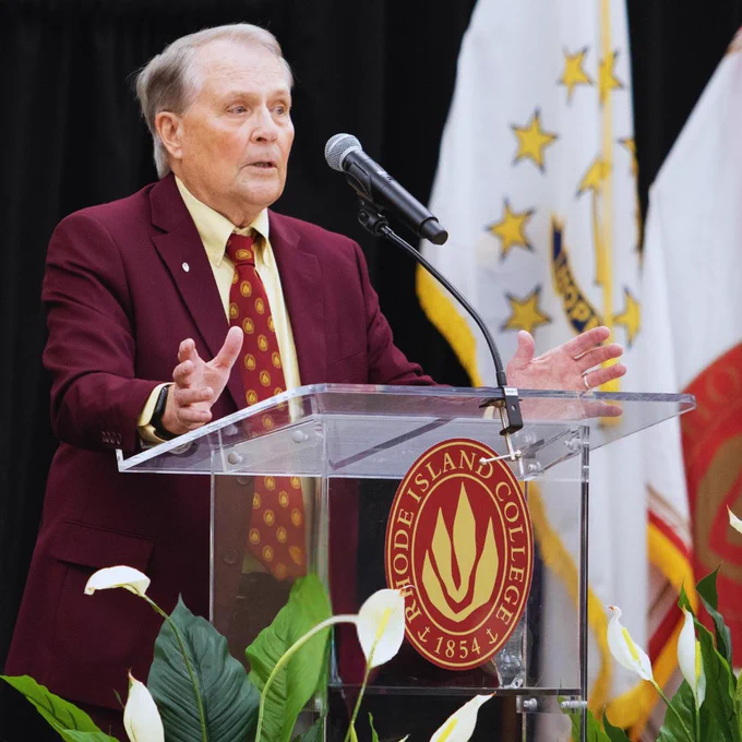 A person speaking at a podium with the Rhode Island College logo. The backdrop features flags, including one with stars. Lush greenery with white flowers decorates the foreground.