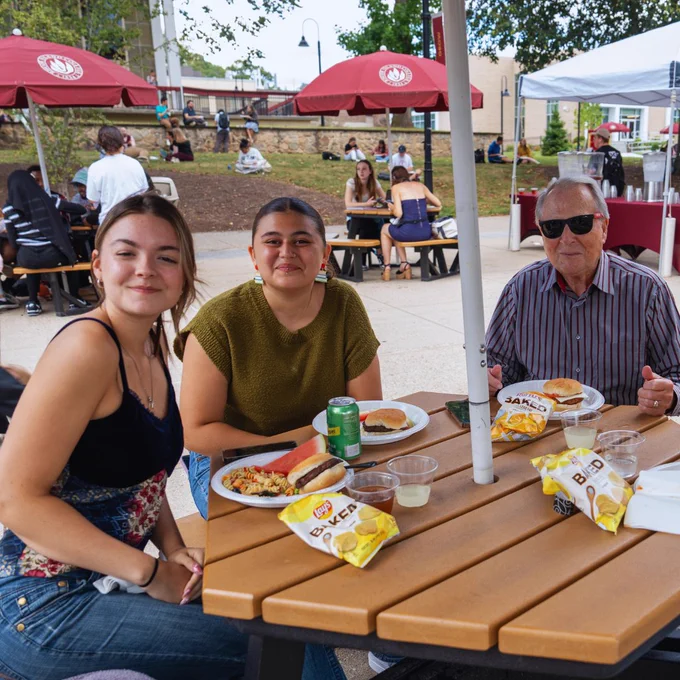 A group of three people sitting at an outdoor picnic table enjoying a meal with plates of food and bottles of drinks. Bags of Lay's chips are on the table. Other people and red umbrellas are visible in the background.
