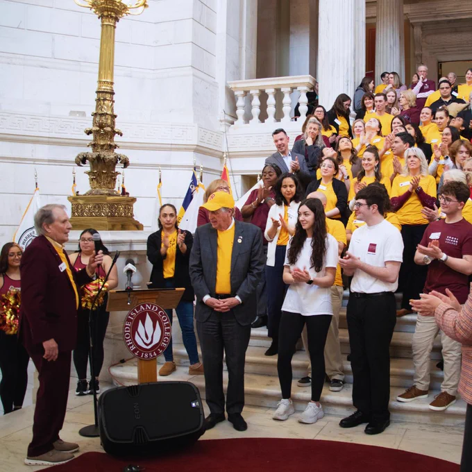 A group of people is gathered inside a grand building with marble floors and walls. Many are wearing yellow and maroon shirts with "Rhode Island College" logos. A large brass decorative fixture is visible. Some individuals are standing on stairs, applauding a person speaking at a podium with "Rhode Island College" inscribed on it. The mood is celebratory.