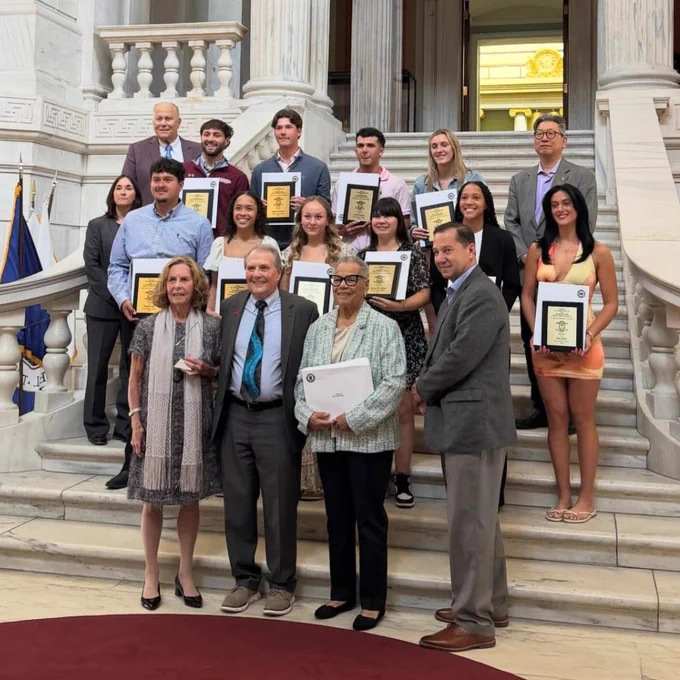 A group of people standing on a grand staircase inside a marble building, holding certificates of recognition. Some individuals are dressed in formal attire, and others in business casual. There is an American flag on one side.