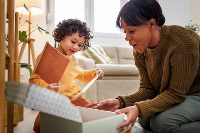 A woman and a child in a living room excitedly opening a shoebox, with the child holding a wooden item and paper.