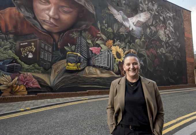 Woman stood next to a mural, painted on a wall, with flowers, a tram, a young person, buildings and nature. 