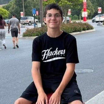 Talon is seated on a large red ball in an outdoor setting, wearing a black "Hackers" shirt and shorts. In the background, there are trees and a few parked cars. Other people are walking nearby.
