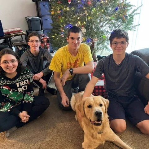 A group of four people sitting on the floor in front of a decorated Christmas tree with colorful lights. A smiling golden retriever is lying in front of them. Wrapped gifts are visible under the tree. 