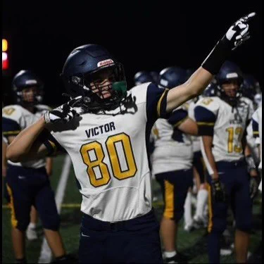Talon is in his #80 Victor football jersey and pads on the sideline at one of his football games.