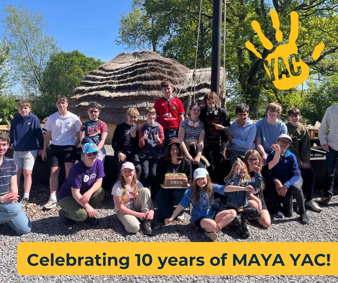 A group of children and adults gathered in front of a reconstructed Anglo-Saxon house on a sunny day. Text at the bottom says 'Celebrating 10 years of MAYA YAC!'