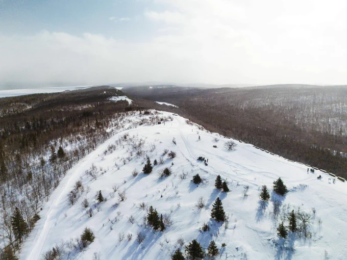 Aerial view of a snowy ridge with scattered pine trees and a winding path through a vast winter forest landscape.