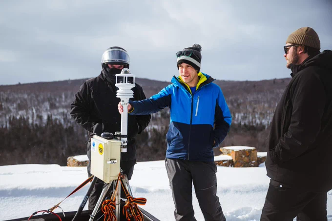 Three Michigan Tech researchers in winter clothing operate a weather monitoring device on a snowy landscape with forested hills in the background.