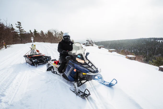 Person riding a snowmobile on a snowy trail pulling a sled with equipment in a winter forest landscape.
