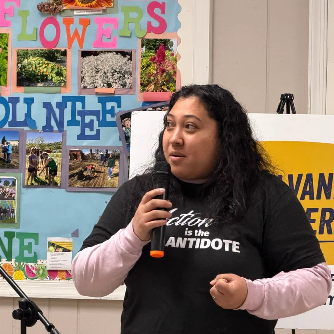 A young latina speaks passionately into a microphone, her t-shirt reads “action is the antidote.” 
