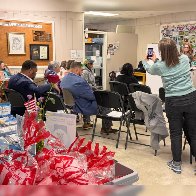 In the foreground is a bucket of colorfully packaged cotton candy, and behind that are people sitting in a community center room. A woman takes a picture with her camera of Northampton County Executive Tara Zrinski speaking.  
