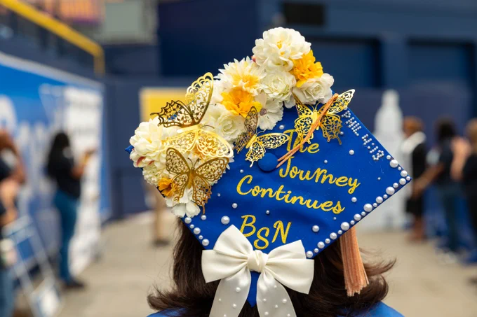 A decorated graduation cap adorned with white flowers, yellow butterflies, and pearl accents is featured. The cap has a white bow at the bottom and displays the message "The Journey Continues BSN" in yellow lettering.