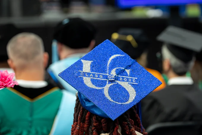 A person in a graduation cap sits among a group of people in academic gowns. The cap is blue with glitter and displays "US Class of 2026.