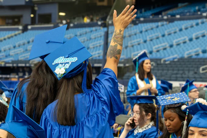 A group of graduates in blue caps and gowns sit in a stadium. The focus is on one graduate with a raised, tattooed arm, whose cap reads "Mama did it!" Other graduates, some with decorated caps, are seated nearby.
