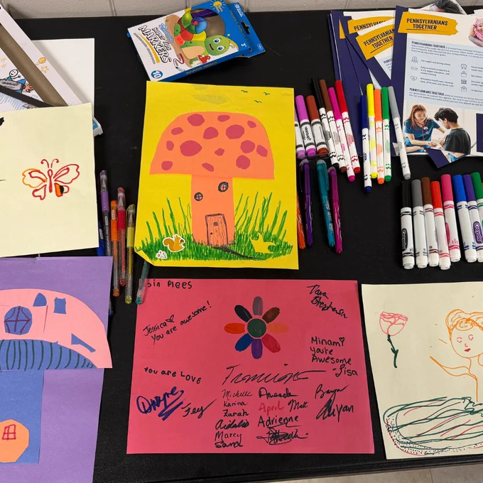 A photo of children’s illustrations on colorful paper, in the top corner is a stack of stickers and papers from Pennsylvanians Together. 
