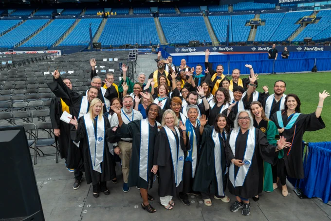 A group of people wearing graduation gowns and sashes stand together on a stadium field, smiling and waving. They are in a fun, celebratory pose with empty seats in the background.