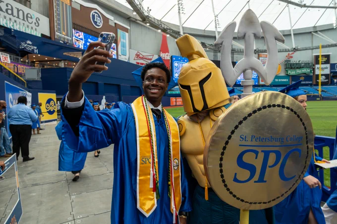 A graduate in a blue cap and gown takes a selfie with SPC mascot Titus, who is wearing a helmet and holding a shield with "SPC" written on it. The scene is inside a stadium, with Tampa Bay Rays logos visible in the background.