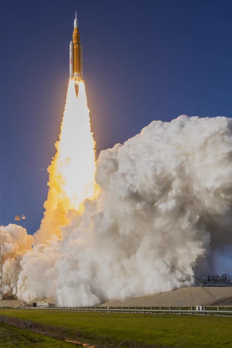 An image of the launch of the Artemis II mission. The SLS (Space Launch System) rocket with the Orion spacecraft is lifting off into the blue sky with a dramatic trail of flames underneath it, morphing into billowing clouds. Credit: NASA/Jim Ross