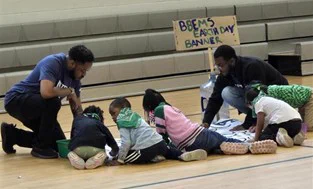 Children and adults collaborate on an Earth Day banner in a gym. The mood is focused and creative.