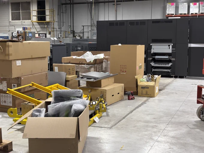 The floor of a commercial print shop mid-transition. Several large dark gray press units are visible in the background, surrounded by open cardboard boxes, bubble-wrapped components, yellow rigging equipment, and various tools scattered across the concrete floor.
