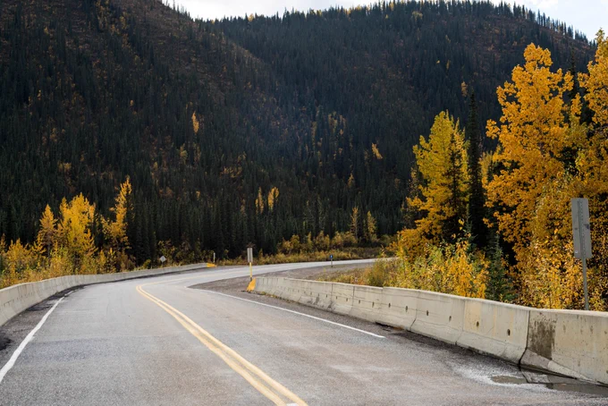 Route de montagne sinueuse bordée de barrières en béton avec des arbres d’automne affichant des feuilles dorées sous un ciel nuageux.