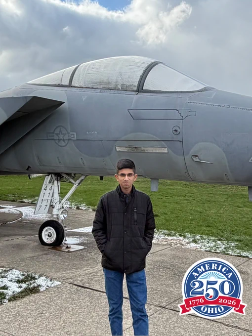 A person stands in front of a military jet on a cloudy day with patches of snow on the ground. The aircraft is gray with military insignia. In the bottom right corner is an "America 250 Ohio 1776-2026" logo.