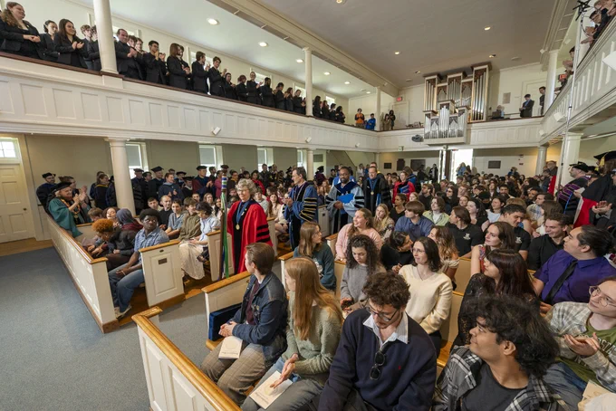 A large gathering in a chapel with rows of seated attendees and others standing in a balcony. Some individuals are dressed in academic robes with varied colors. The setting has wooden pews and a high ceiling, creating a formal atmosphere.