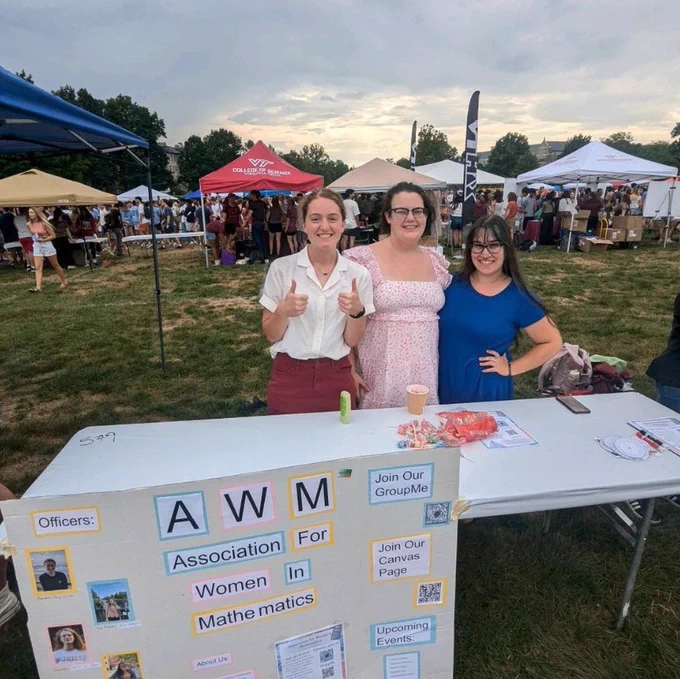 Three women from the Virginia Tech AWM Student Chapter standing behind a table promoting the Association for Women in Mathematics at an outdoor event with tents and attendees in the background.