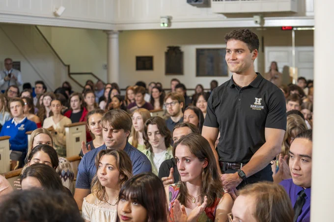 A person standing in a crowded room, surrounded by seated individuals. The standing person is wearing a dark polo shirt with a logo. The room is filled with attentive people seated in rows, clapping. The setting appears to be a formal gathering or ceremony.