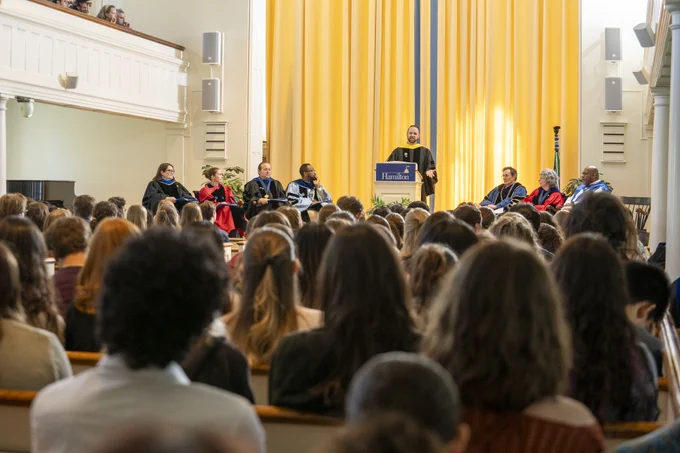 A speaker stands at a podium labeled "Hamilton," addressing a seated audience. Several individuals in academic robes are seated on stage, with yellow curtains in the background. The audience is attentively listening, filling the room's pews.