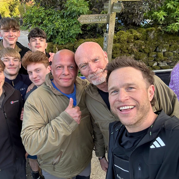 A group of seven people, including singer Olly Murs, pose for a selfie outdoors near a signpost.