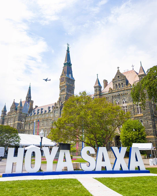 Georgetown University campus scene with large "HOYA SAXA" letters displayed on the grass. Healy Hall's iconic clock tower is visible in the background, and a plane is flying in the sky above.