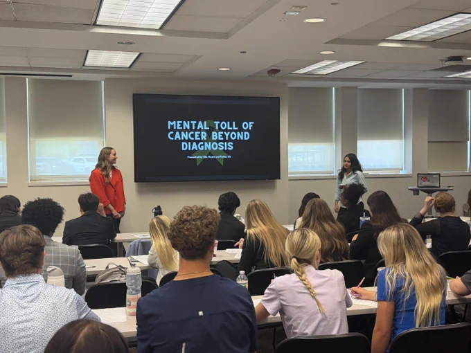 A group of people sitting in a classroom-like setting, listening to a presentation on a large screen that says "Mental Toll of Cancer Beyond Diagnosis".