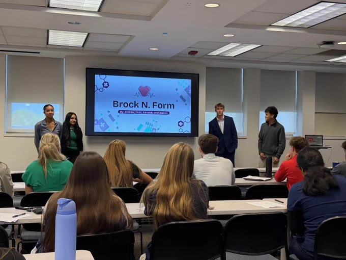 Classroom with students and presenters gathered around a large screen displaying "Brock N. Form" presentation.