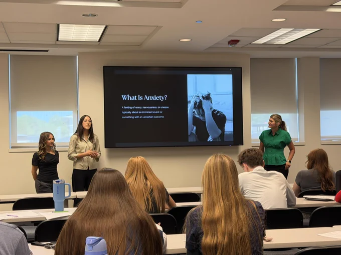 Classroom setting with a presentation on a projection screen showing the text "What is anxiety?". Four women are standing in front of the class and there are students seated at desks.