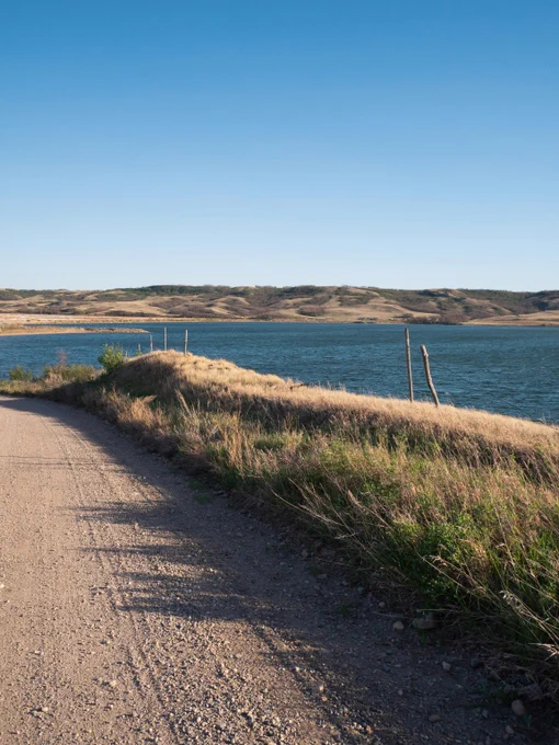 Gravel road curving alongside a calm blue lake with grassy hills under a clear sky.