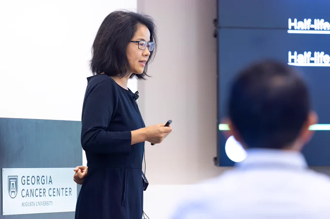 A female scientist, wearing a microphone and holding a remote control for a slide show, presents her scientific findings to a large group of fellow researchers inside a modern lecture hall on the campus of a university.