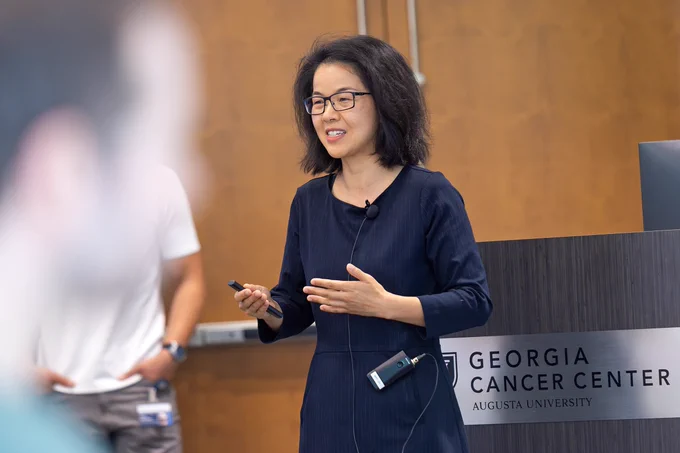 A female scientist, wearing a microphone and holding a remote control for a slide show, presents her scientific findings to a large group of fellow researchers inside a modern lecture hall on the campus of a university.
