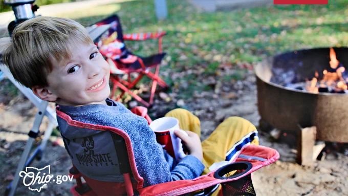 A young boy holds a mug while he sits in a lawn chair next to a campsite fire ring.